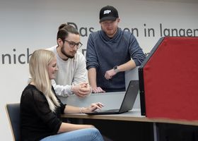 Drei Studierende schauen in der Bibliothek auf den Bildschirm eines Laptops